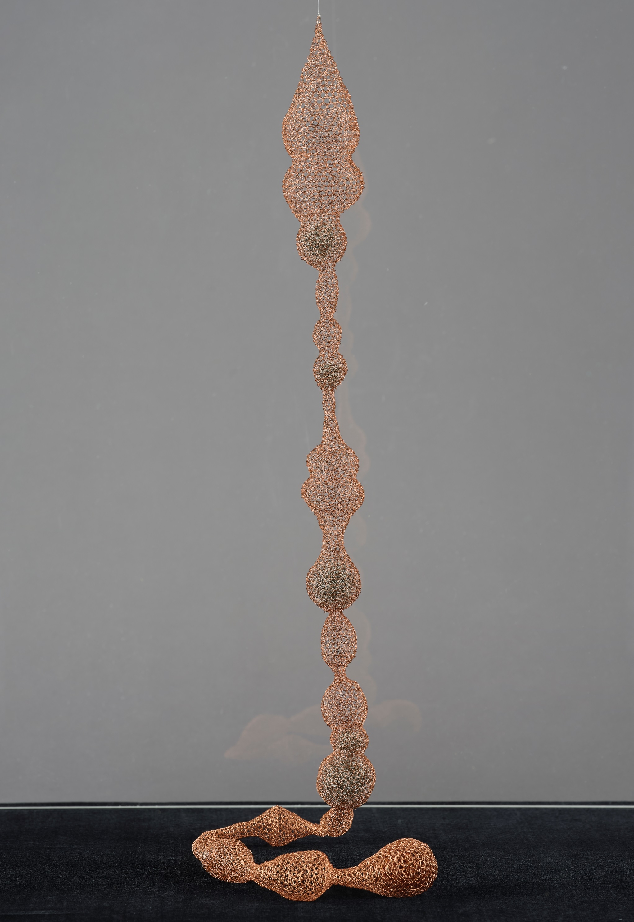 這件作品〈串〉，以金銅色絲線細密的編織出長條的網子，頂端懸吊的垂墜水滴形狀，擬造金屬網子柔軟的樣態。